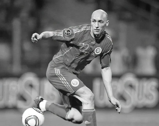 Fekete-fehér fotó egy futballistáról a román válogatott mezében, 22-es számmal, aki labdával a lábánál mozgás közben látható egy stadionban, háttérben elmosódott reklámtáblák.