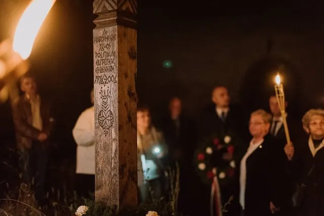 Esti megemlékezés faragott, székely motívumokkal díszített emlékoszlop előtt, körülötte fáklyát tartó emberek és koszorú áll.