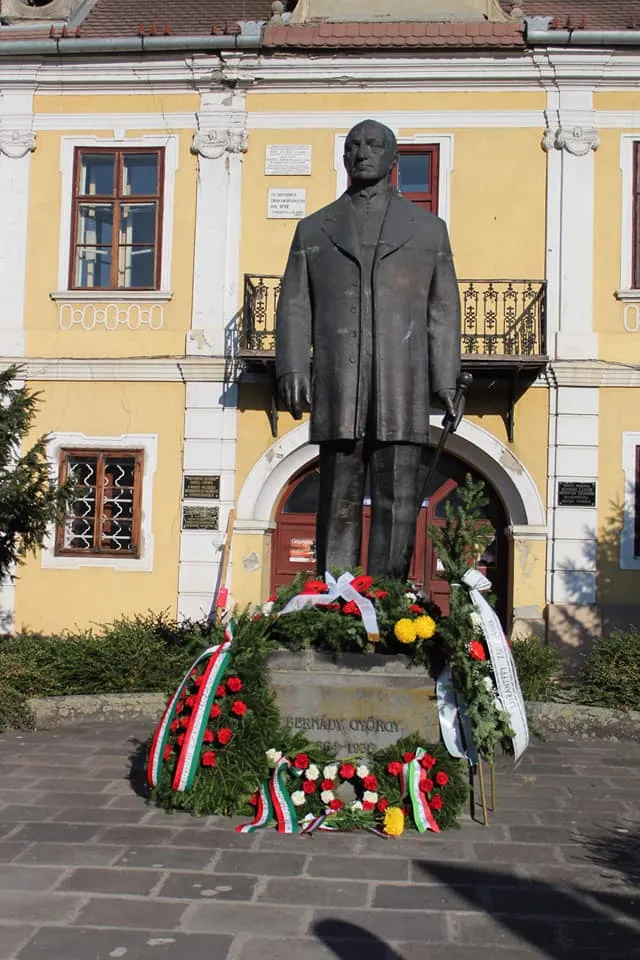 Bernády György szobra egy sárga, historizáló stílusú épület előtt, talapzatánál nemzetiszínű szalagokkal díszített koszorúkkal és fenyőágakkal.