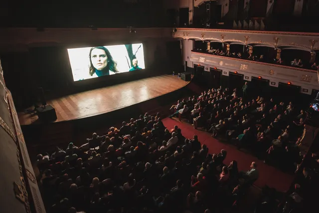 Zsúfolt színházterem felülnézetből, a nézők egy filmvetítést néznek, a nagy kivetítőn két személy közeli portréja látható. A díszes, erkélyes terem a Kultúrpalota nagytermére utal.