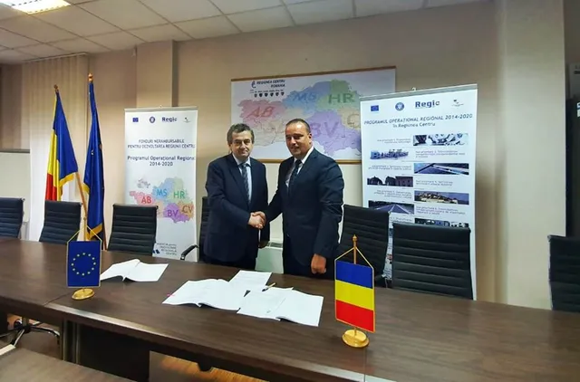 Két öltönyös férfi kezet fog egy tárgyalóteremben, kék asztalon aláírt dokumentumok, mellette román és EU zászlócska, a háttérben Regionális Operatív Program feliratú roll-up bannerek és Románia régiós térképe.