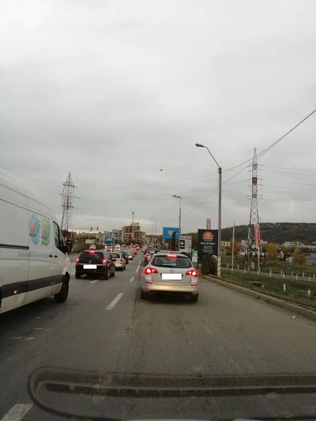 Autós felvétel egy kétsávos útról, ahol hosszan álló autósor torlódik, kilátás felett magasfeszültségű oszlopokkal és domboldallal a háttérben.