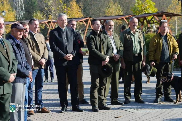 Vadászruhás férfiak csoportja áll sorban egy téren, ősszel, kutyákkal (közöttük egy dobermann), a háttérben fa pergola és fák láthatók.