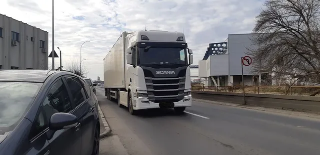 Fehér Scania kamion halad egy úton, mellette parkoló autó látható, a háttérben ipari épületek és egy behajtani tilos tábla.