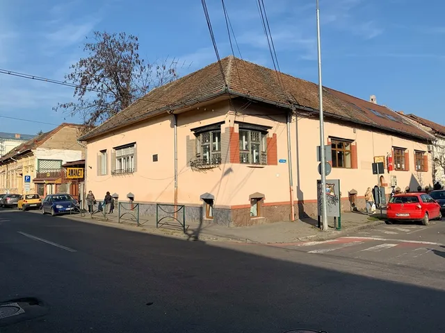 Utcasarki régi, egyszintes sárga polgárház magas cseréptetővel, zsalugáteres ablakokkal, előtte parkoló autók és néhány gyalogos, mellette egy „Amanet” felirású üzlethelyiség.