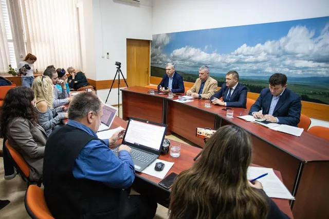 Sajtótájékoztató egy tanácsteremben, ahol négy férfi ül az asztalnál egy nagy tájképfestmény előtt, körülöttük újságírók és jelenlévők ülnek, jegyzetelnek és laptopokkal dolgoznak.