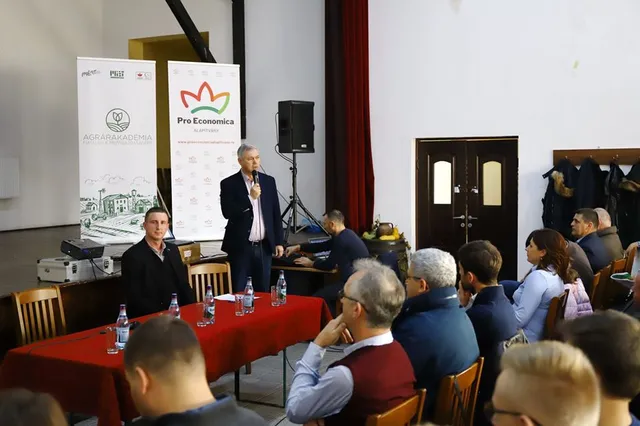 Előadóterem, ahol egy férfi mikrofonnal a kezében áll és beszél egy piros terítővel borított asztal mellett, míg a hallgatóság ülve figyeli. A háttérben roll-up bannerek láthatók 'Agrárakadémia' és 'Pro Economica' felirattal.