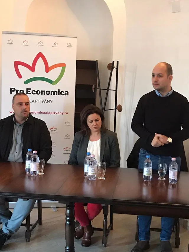 Két férfi és egy nő ül, illetve az egyik férfi áll egy asztalnál, mögöttük Pro Economica Alapítvány feliratú roll-up banner látható egy boltíves teremben, az asztalon vizesüvegek és poharak.