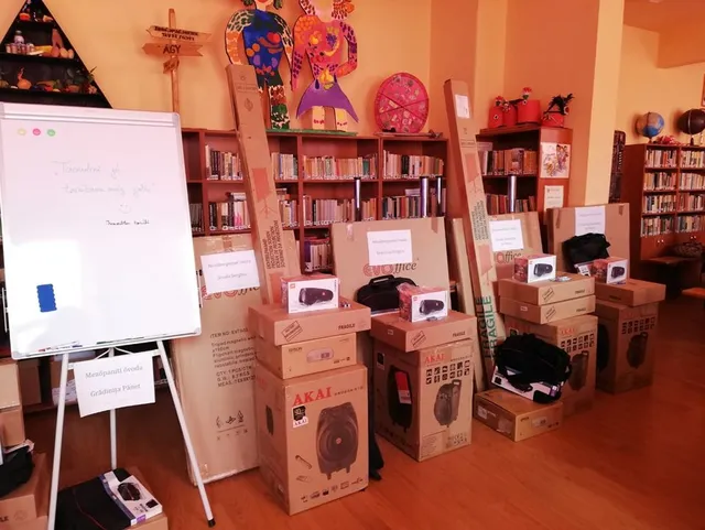 Könyvtárterem polcokkal, előtérben dobozolt digitális eszközök (hangszórók, projektorok, táskák) egymásra pakolva, mellette flipchart tábla és felirat 'Mezőpanit Iskola Grădeşti Pinet'.