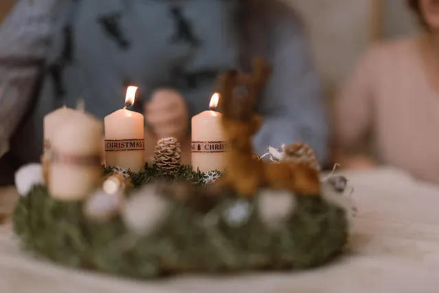 Adventi koszorú fenyőágból, égő és el nem égett fehér gyertyákkal, fenyőtobozokkal és apró dísztárgyakkal, háttérben elmosódottan két személy ül az asztalnál.