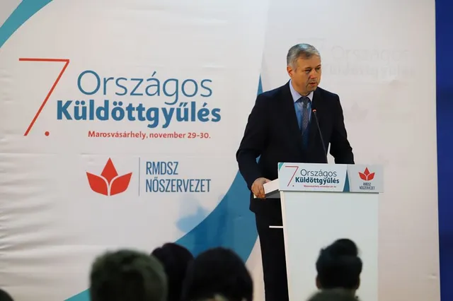 Öltönyt viselő férfi beszél egy pulpituson, mögötte az RMDSZ Nőszervezet 7. Országos Küldöttgyűlésének feliratos háttértáblája látható, előtérben ülő közönség tarkója.