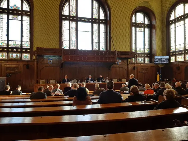 Nagy, díszes teremben tartott tanácsülés látható, díszes faburkolattal, magas boltíves ablakokkal és festett mennyezettel. Elöl az elnökség asztalánál ülnek a tisztségviselők, a padsorokban tanácstagok foglalnak helyet, egy férfi éppen felállva beszél.