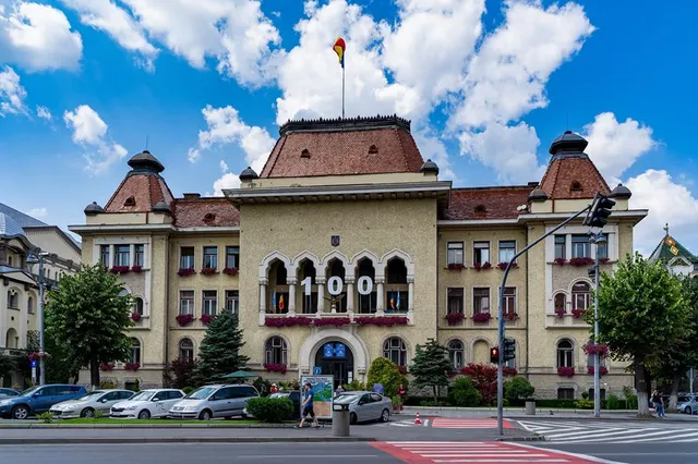 A marosvásárhelyi polgármesteri hivatal épülete napsütésben, homlokzatán '100' feliratú dísszel, előtte parkoló autók és néhány sétáló ember.