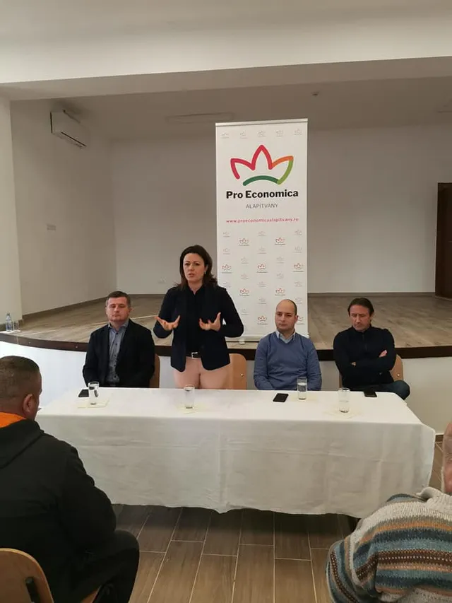 Négy ember ül egy asztalnál egy teremben, középen egy nő áll fel és beszél kezeivel gesztikulálva. Mögöttük a Pro Economica Alapítvány roll-up bannere látható, előttük hallgatóság ül.