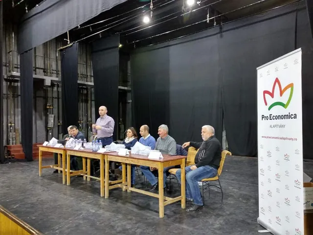 Színpadszerű teremben, sötét háttérfüggönyök előtt egy hosszú asztal mögött öt férfi és egy nő ül, egyikük áll és beszél mikrofonba, mellettük a Pro Economica Alapítvány rollup bannere látható.
