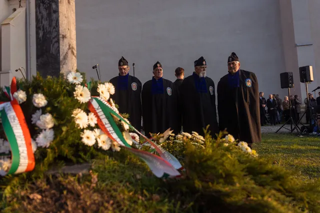 Négy díszegyenruhás, sapkás férfi áll sorban egy megemlékezésen, előttük nemzeti színű szalagos koszorúk és fehér virágcsokrok, mögöttük egy világos épület fala, oldalt hangosító berendezés és nézők állnak.