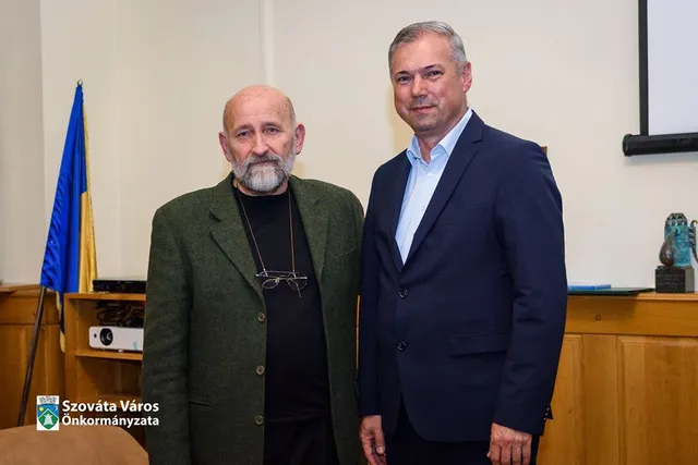 Két férfi áll egymás mellett egy teremben, mögöttük ukrán-kék zászló és fa berendezésű tanácsterem részlete látszik. A bal oldali idősebb férfi zöld kabátot visel, szemüveg lóg a nyakában, a jobb oldali fiatalabb férfi sötétkék öltönyt és világoskék inget hord.