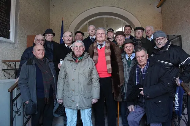 Idős férfiak nagyobb csoportja pózol csoportképen egy épület boltíves bejárata előtt, télikabátban, néhányan botra támaszkodva.
