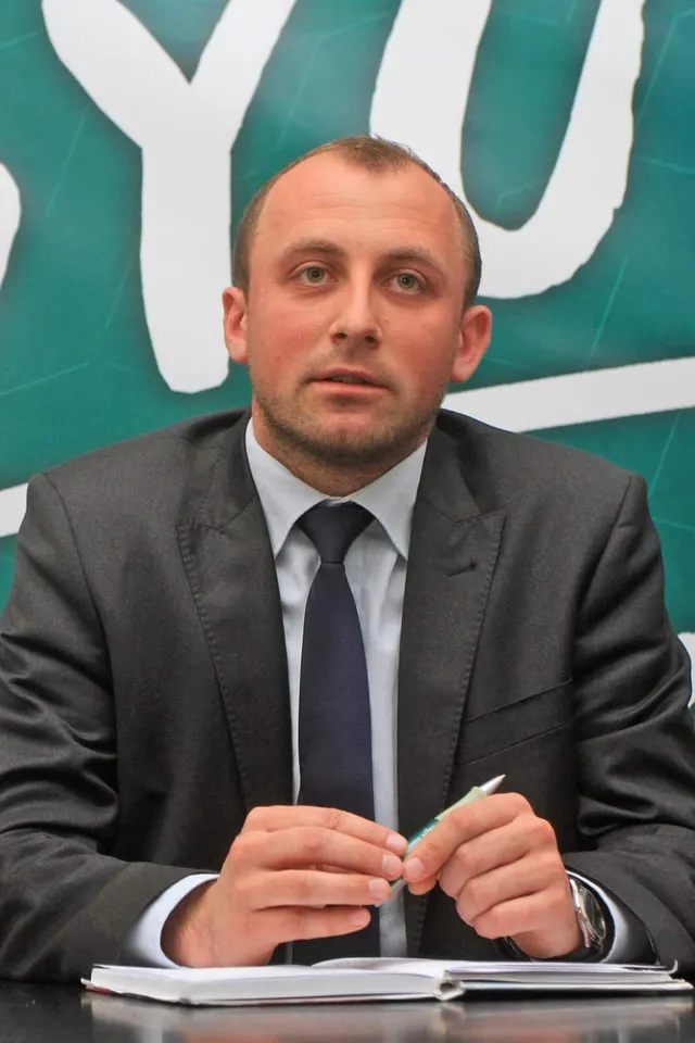 Sötét öltönyt, világoskék inget és sötétkék nyakkendőt viselő férfi ül egy asztalnál, kezében tollat tart, előtte nyitott jegyzetfüzet, mögötte zöld hátterű molinó felirattal.