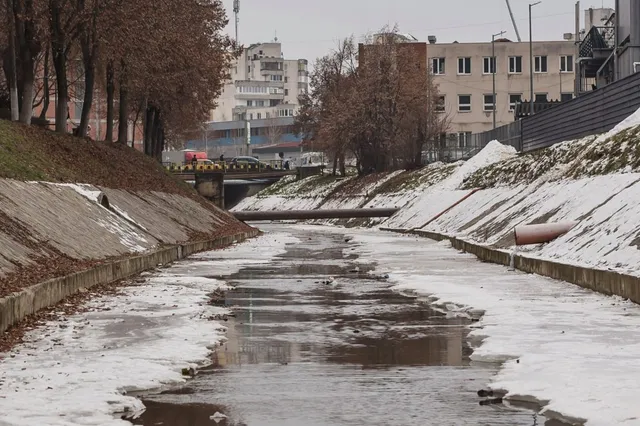 Betonozott mederrel szegélyezett patak télen, jégfoltokkal és hóval a partokon, háttérben egy híd és városi épületek.
