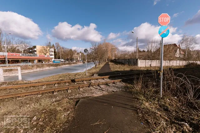 Vasúti sín menti aszfaltozott gyalog- és kerékpárút látható, mellette STOP tábla és kerékpáros jelzőtábla áll. A háttérben úttest, buszmegálló, üzletsor (Darina Supermarket) és lakóépületek láthatók, kék égen felhőkkel.