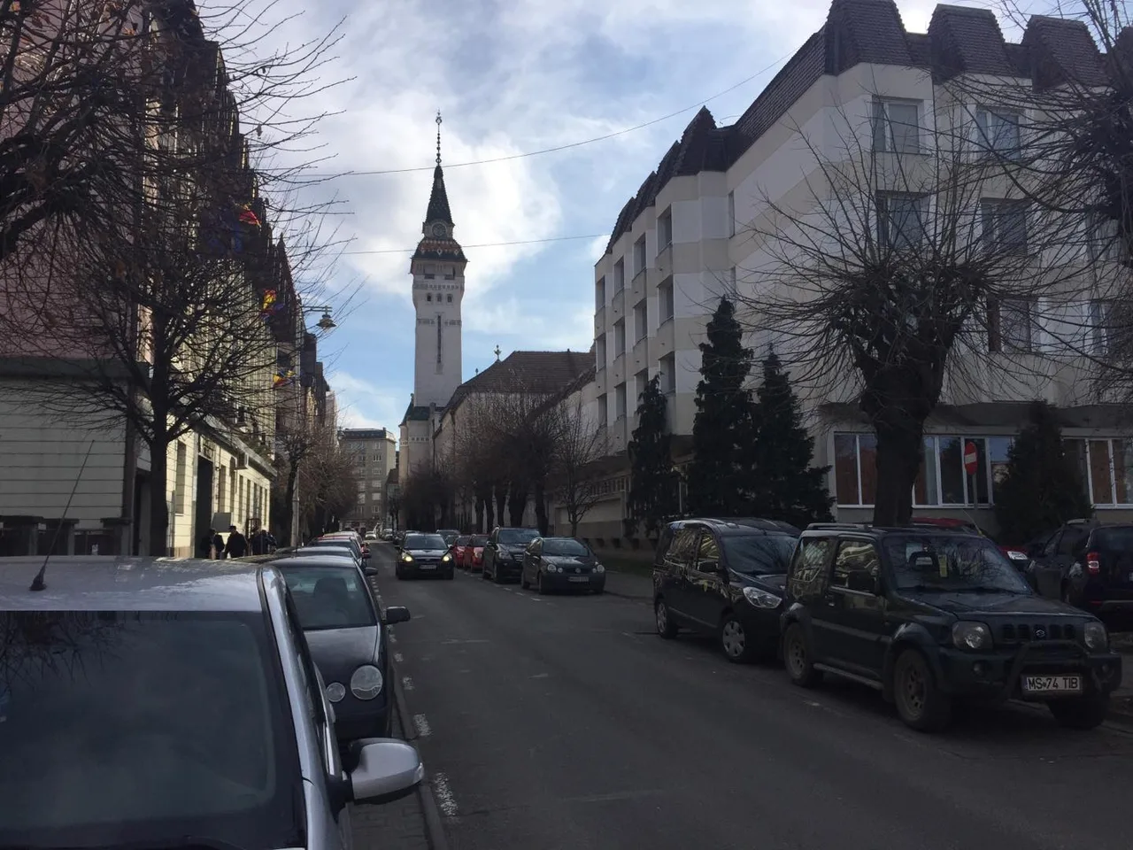 Utcakép Marosvásárhelyen, kétoldalt parkoló autókkal, háttérben a Városháza (Prefektúra) jellegzetes órástornya látszik, tél végi, lombtalan fákkal szegélyezett úton.