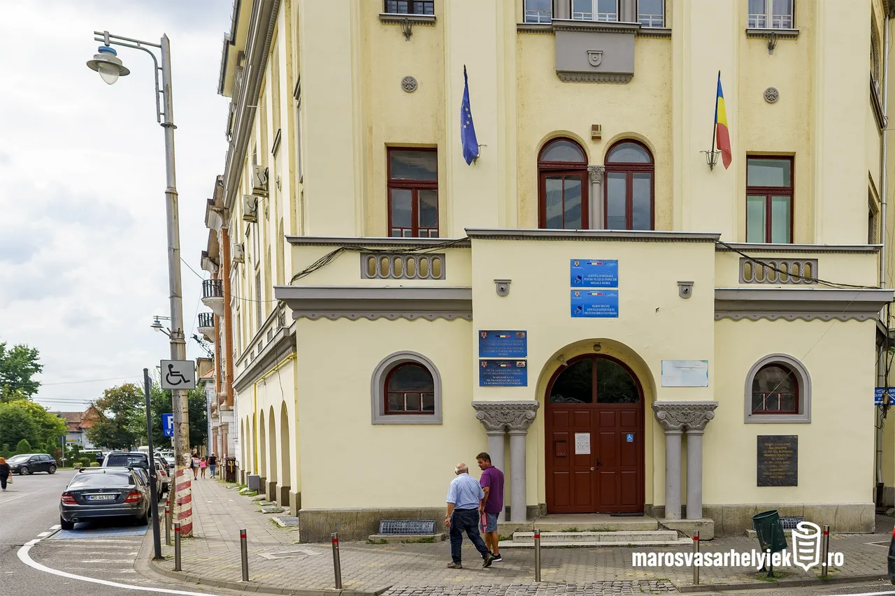 Sárga, klasszikus stílusú intézményi épület bejárata, amelyen EU- és román zászló, illetve kék-fehér intézményi táblák láthatók. Az épület előtt a járdán két férfi beszélget, mögöttük parkoló autók.