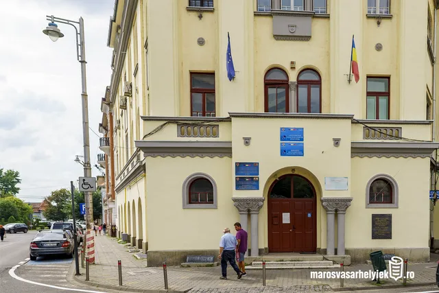 Sárga, klasszikus stílusú intézményi épület bejárata, amelyen EU- és román zászló, illetve kék-fehér intézményi táblák láthatók. Az épület előtt a járdán két férfi beszélget, mögöttük parkoló autók.
