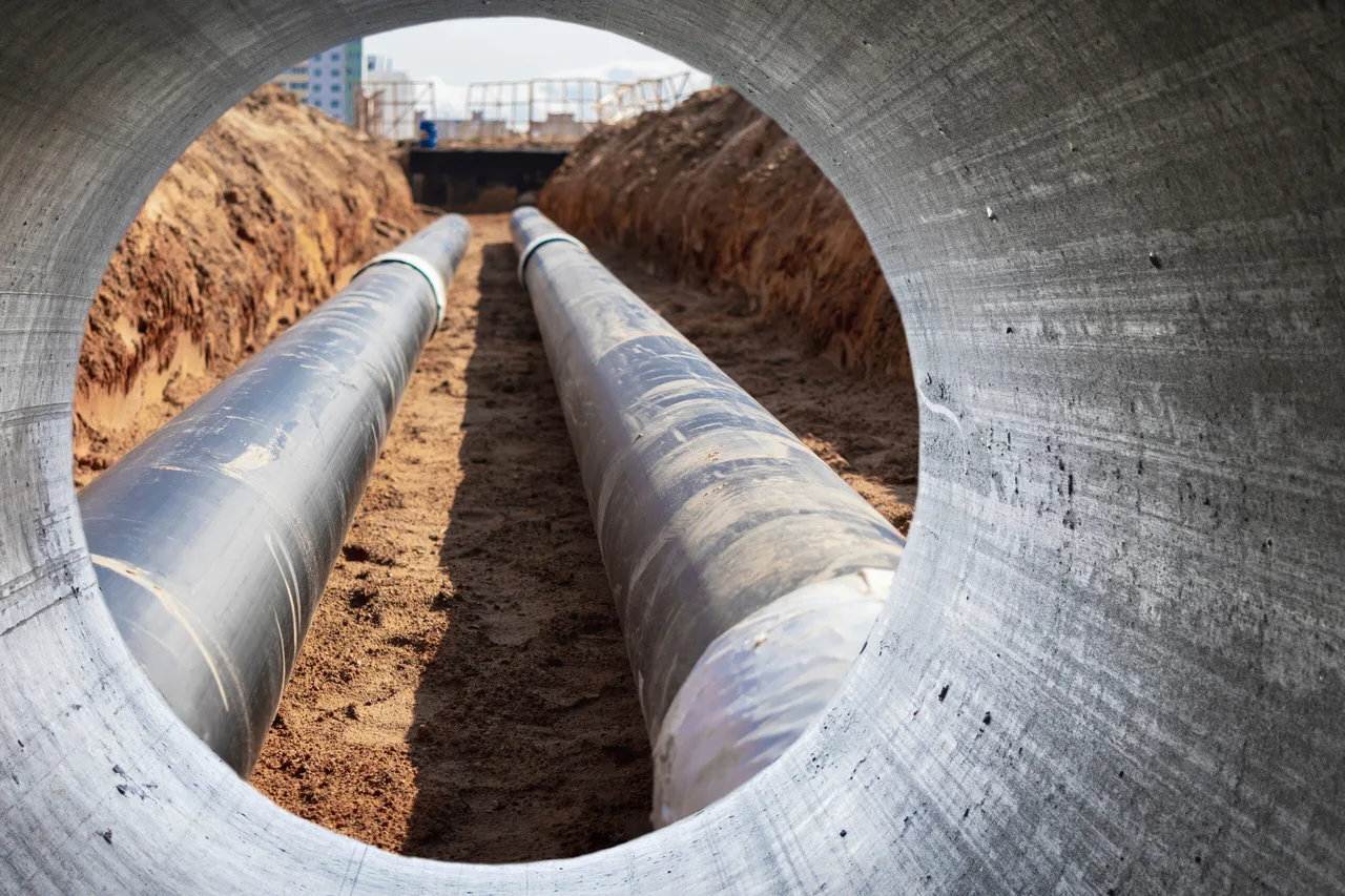 Betonelem nyílásán át egy kiásott munkaárok látható, benne két nagy átmérőjű, szigeteléssel bevont csővezetékkel; háttérben földkupacok és egy építkezés alatt álló magas épület körvonala.