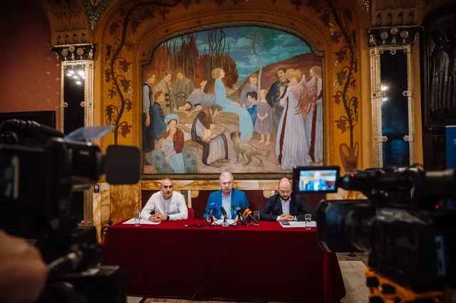 Sajtótájékoztató a Kultúrpalota egyik díszes termében, egy nagy freskó előtt: három férfi ül vörös terítővel fedett asztal mögött, mikrofonokkal, előttük kamerák rögzítik a beszélgetést.