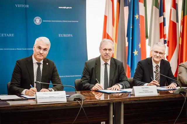 Három öltönyös férfi ül egy asztalnál, dokumentumokat írnak alá, mögöttük kék molinó a Beruházási és Európai Projektek Minisztériumának feliratával, valamint uniós és egyéb országok zászlói sorakoznak.