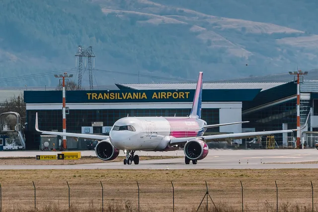 Rózsaszín-fehér Wizz Air repülőgép áll a futópályán a Marosvásárhelyi Transilvania Nemzetközi Repülőtér terminálja előtt, háttérben hegyek láthatók.