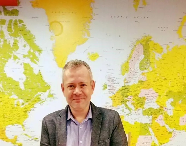 Középkorú férfi áll egy nagy méretű, színes világtérkép előtt, szürke zakóban és világoskék ingben, mosolyogva a kamerába néz.
