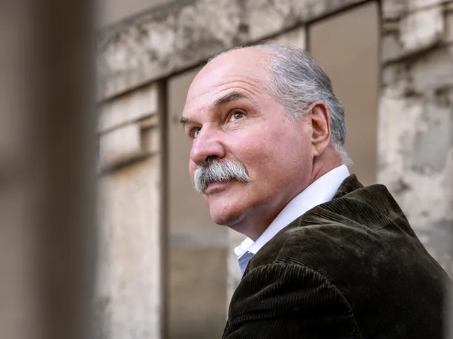 Idősebb, ősz hajú és bajszú férfi profilból, kordbársony zakóban és fehér ingben áll egy épület előtt, oldalra és felfelé tekint.