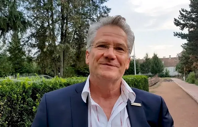 Ősz hajú, szemüveges férfi mosolyog a kamerába, sötétkék zakóban és mintás ingben, háttérben fák, bokrok és egy régi épület látszik alkonyati fényben.