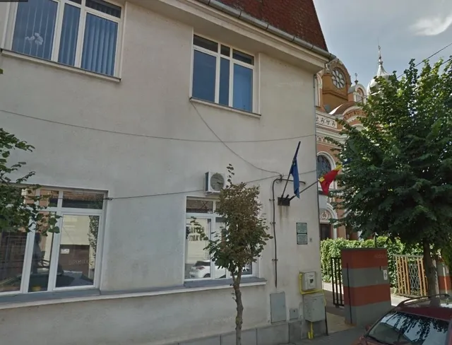 Egy sarki épület fehér homlokzata látható, ablakokkal, klímaberendezéssel és kihelyezett EU- és román zászlóval; a háttérben egy templom tornya és órája látszik.