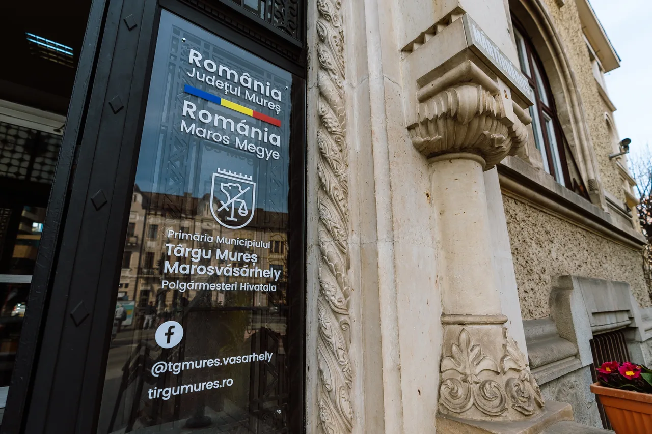 Üvegtáblás bejárati tábla, rajta a Marosvásárhelyi Polgármesteri Hivatal kétnyelvű felirata, Maros megye címere és elérhetőségei, mellette egy díszesen faragott kőoszlop és a városháza homlokzata.