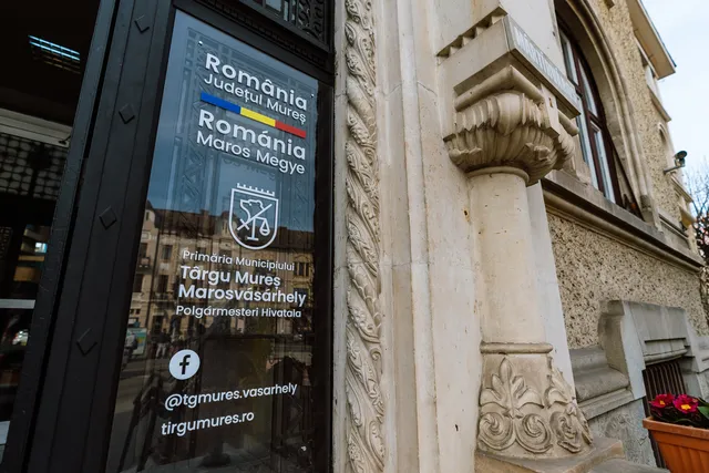 Üvegtáblás bejárati tábla, rajta a Marosvásárhelyi Polgármesteri Hivatal kétnyelvű felirata, Maros megye címere és elérhetőségei, mellette egy díszesen faragott kőoszlop és a városháza homlokzata.