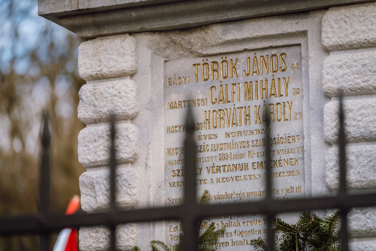 Kőoszlop felirata a Székely Vértanúk emlékművén, aranybetűkkel Török János, Csálfi Mihály és Horváth Károly nevével, akiket 1854. március 10-én végeztek ki, előtte vaskerítés és fenyőág díszítés.