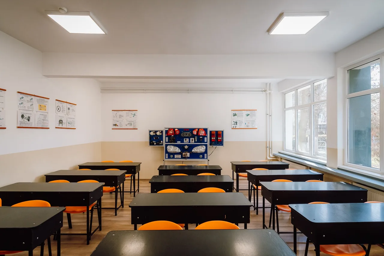Üres, felújított tanterem padokkal és narancssárga székekkel, a hátsó falon autóalkatrészeket (fényszórókat, műszerfalat) bemutató oktatási panel, oldalt nagy ablakok.