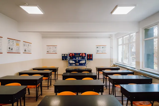 Üres, felújított tanterem padokkal és narancssárga székekkel, a hátsó falon autóalkatrészeket (fényszórókat, műszerfalat) bemutató oktatási panel, oldalt nagy ablakok.