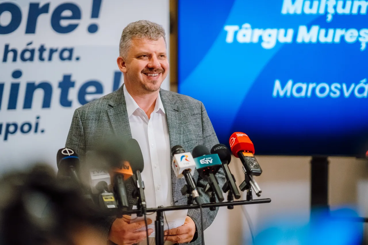 Öltönyt viselő, mosolygó férfi sajtótájékoztatón beszél, előtte számos mikrofon látható a tudósító stúdiók logóival, mögötte kék háttérvetítés Marosvásárhely feliratokkal.