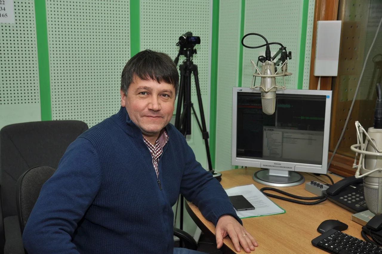 Egy férfi ül rádiós stúdióban íróasztal mellett, mögötte mikrofon, monitor hangszerkesztő programmal, telefon és állványos kamera látható.