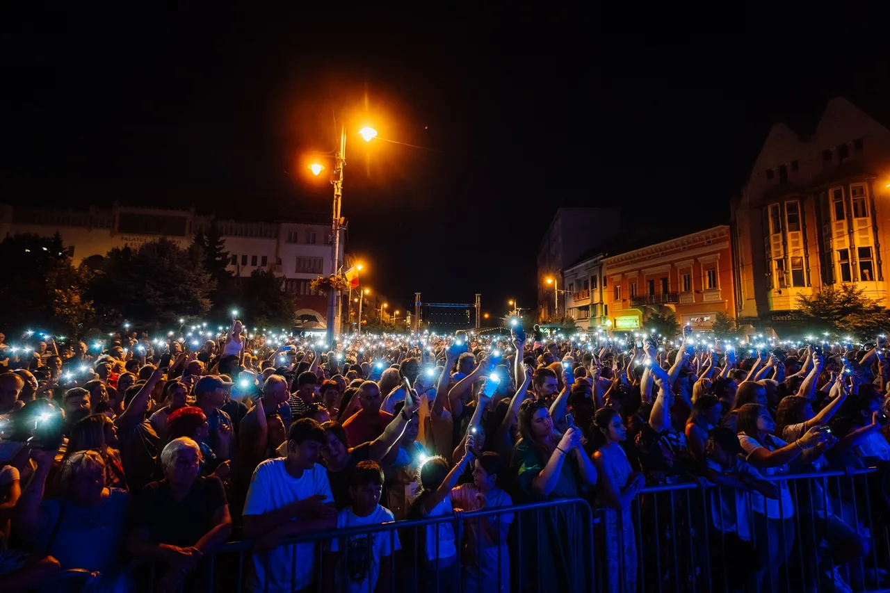 Éjszakai tömeg gyűlt össze egy utcai rendezvényen, sok néző emeli fel telefonját, a bekapcsolt lámpák fénypontokat alkotnak a sötétben, mögöttük megvilágított városi épületek látszanak.