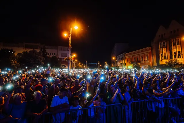 Éjszakai tömeg gyűlt össze egy utcai rendezvényen, sok néző emeli fel telefonját, a bekapcsolt lámpák fénypontokat alkotnak a sötétben, mögöttük megvilágított városi épületek látszanak.