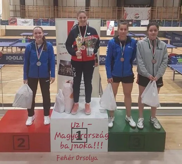 Négy fiatal női asztaliteniszező áll dobogón, érmekkel a nyakukban, sportcsarnokban. A győztes (1. helyezett) trófeát és virágcsokrot tart, mellette rátett felirat: „UZI – Magyarország bajnoka!!! Fehér Orsolya”.