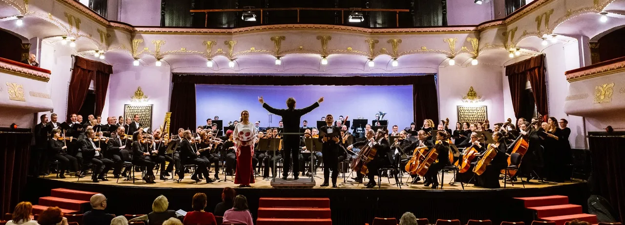 Zenekari koncert egy díszes, erkélyes koncertteremben: karmester vezényel, mellette énekesnő és férfi szólista áll a színpad elején, körülöttük vonós- és fúvós zenekar ül, előtérben nézőtér látható.