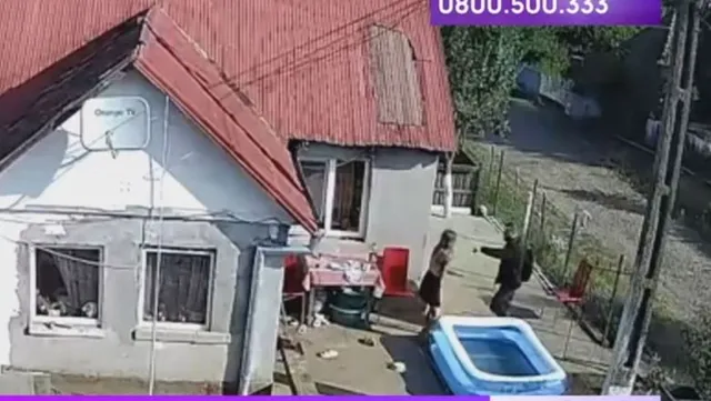 Biztonsági kamera felvétele egy udvarról: piros tetős ház előtt két személy áll, egy nő és egy férfi, közöttük feszült interakció látszik, a földön gyerekmedence és kerti bútor található.