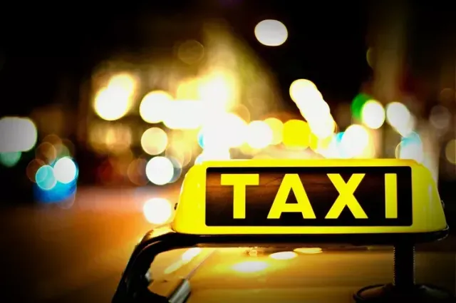 Éjszakai felvétel egy taxi tetején lévő, sárga fényű, megvilágított „TAXI” feliratú jelzőtábláról, a háttérben elmosódott fények és fényfoltok látszanak.