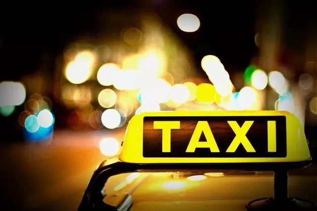 Éjszakai felvétel egy taxi tetején lévő, sárga fényű, megvilágított „TAXI” feliratú jelzőtábláról, a háttérben elmosódott fények és fényfoltok látszanak.